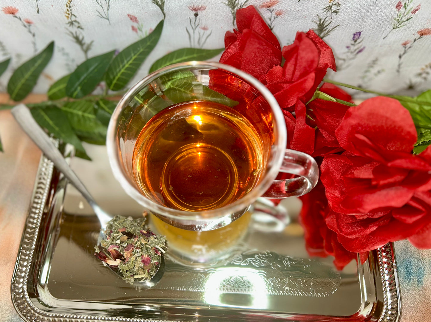 Glass tea cup of Calming herbal tea alongside a teaspoon with dried tea blend of tulsi, rose, and rooibos for stress relief and emotional balance